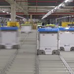 Orange warehouse robots carrying tall shelving units across a busy fulfilment centre floor.