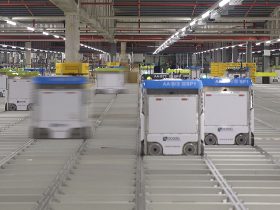 Orange warehouse robots carrying tall shelving units across a busy fulfilment centre floor.